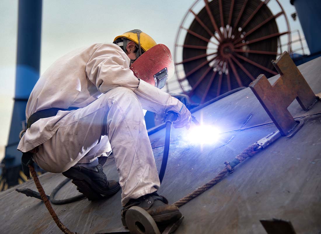 Marine Contractor Insurance - A Welder Suspended With Cables and Working on a Large Ship in a Shipyard in the Evening