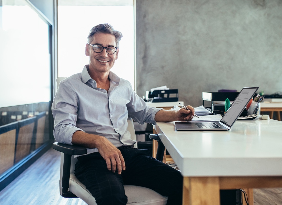 Business Insurance - Businessman Smiling at Camera While Sitting at a Desk in a Modern Office